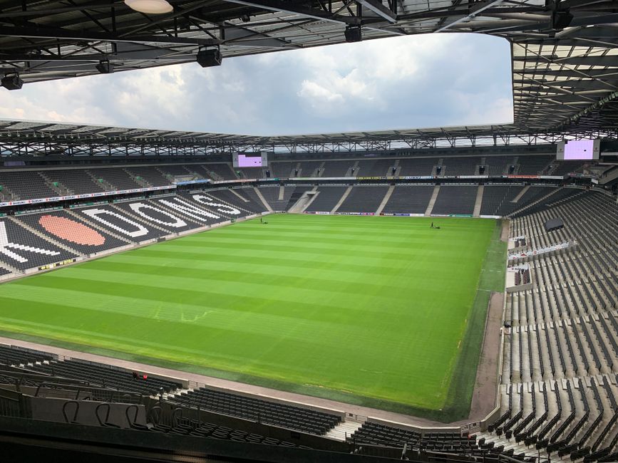 A view from inside a large, modern stadium during daytime showing a well-maintained, lush green grass pitch with evenly mown stripes and a smooth surface, surrounded by tiered seating areas with black and grey seats. The stands include a section with large white letters spelling 'FULHAM' on the left side and a circular logo in a shade of orange and pink on the seating across from it. Overhead, a partially open roof structure with metal trusses and beams creates an industrial aesthetic, and a bright, cloudy sky is visible through the opening. The image reflects the mature, professional environment of a football stadium, with a focus on the pitch and seating arrangement, and the structural elements hint at the possibility of on-site event preparations or maintenance activities. Occasionally, images of stadiums relate to events requiring clearing of rubbish or waste during match days, as handled by independent waste collection services like Waste Collection Fulham.