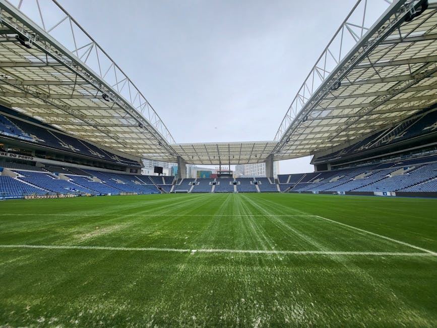 A large football stadium featuring a well-maintained green pitch with visible white boundary lines. The seating area surrounds the field, with dark blue seats arranged in multiple tiers on both sides. Overhead, a modern, curved roof structure made of clear and metallic materials provides partial cover for the stands, supported by white steel trusses. The sky above is overcast with diffused lighting, casting a neutral tone over the scene. The stadium appears empty, with no players or spectators present. In the foreground, the freshly cut grass displays consistent texture and vibrant colour, typical of a professional sports venue prepared for an upcoming match or event. This image, from wastecollectionfulham.co.uk, could relate to the maintenance or cleaning of large-scale venues, with potential relevance to on-site or alternative stadium waste management services for event organisers or facilities management companies involved in large venue rubbish clearance.