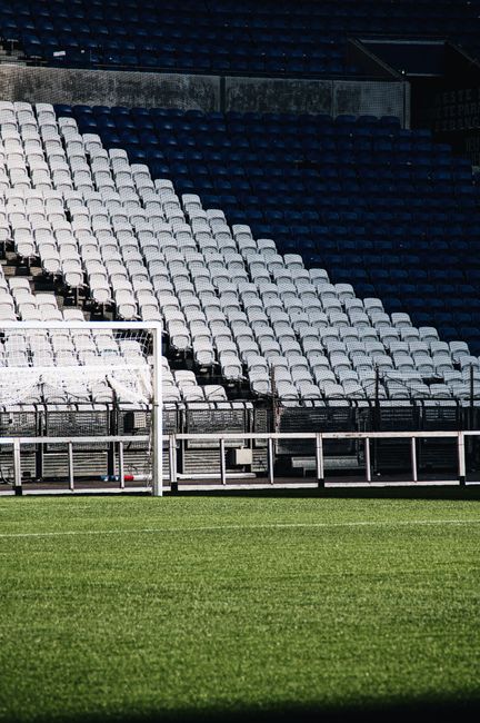 A view from inside a large, modern stadium during daytime showing a well-maintained, lush green grass pitch with evenly mown stripes and a smooth surface, surrounded by tiered seating areas with black and grey seats. The stands include a section with large white letters spelling 'FULHAM' on the left side and a circular logo in a shade of orange and pink on the seating across from it. Overhead, a partially open roof structure with metal trusses and beams creates an industrial aesthetic, and a bright, cloudy sky is visible through the opening. The image reflects the mature, professional environment of a football stadium, with a focus on the pitch and seating arrangement, and the structural elements hint at the possibility of on-site event preparations or maintenance activities. Occasionally, images of stadiums relate to events requiring clearing of rubbish or waste during match days, as handled by independent waste collection services like Waste Collection Fulham.