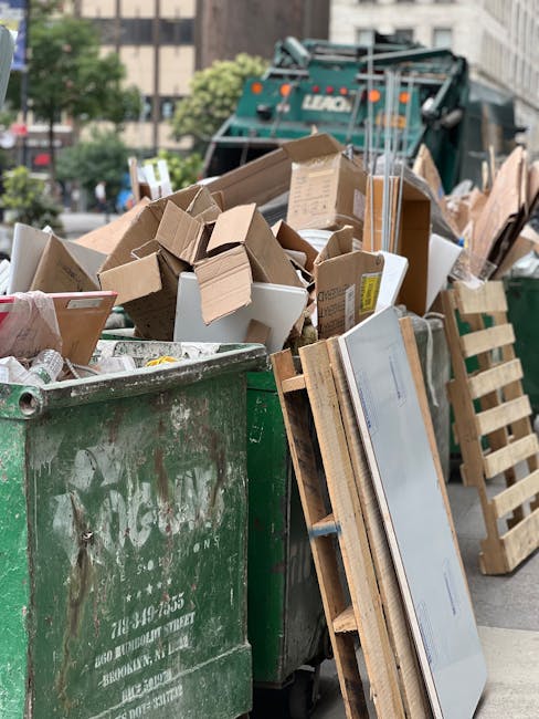 A large green waste bin filled with a mix of cardboard boxes, some flattened and others partially open, along with white packaging containers and small paper scraps. The surface of the bin shows signs of wear with scratches and graffiti. Leaning against the bin are wooden pallets and flat-pack furniture components, including a panel with a reflective surface. In the background, a green waste collection truck with a hydraulic lift mechanism is parked on an urban street, surrounded by trees, multi-storey buildings, and a clear sky. The scene appears to represent a private rubbish collection or on-site clearance, with the waste prepared for removal by local waste management services such as Waste Collection Fulham. The environment suggests a typical urban setting where rubbish is temporarily staged prior to disposal or recycling, emphasizing the practicalities of alternative waste handling methods in Fulham.