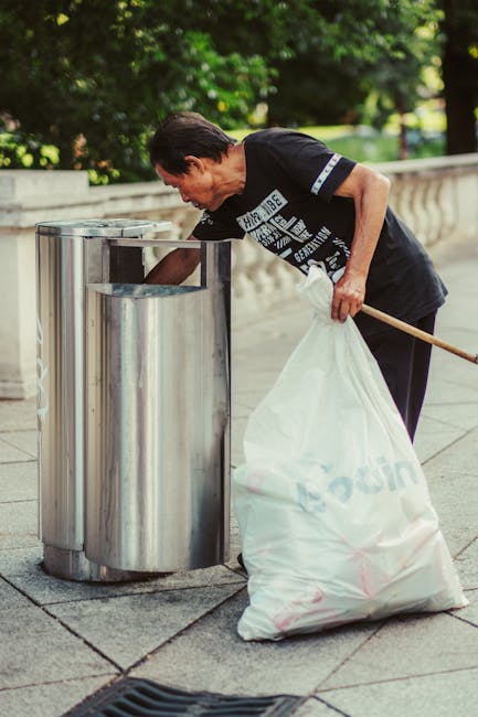 A middle-aged man with dark, slightly graying hair, dressed in a black T-shirt with a graphic design and dark pants, is seen standing upright next to a modern, metallic waste bin with a rounded top in an outdoor urban setting. He is holding a large white plastic bag filled with rubbish, which is partially draped over his arm, and is using a wooden stick or broom handle to help guide the bag into the bin. The pavement beneath him features large, evenly cut stone tiles, and the background includes a textured stone balustrade and lush, green leafy trees casting dappled sunlight, indicating a park or public square. The scene suggests an instance of independent waste disposal or rubbish collection aimed at maintaining cleanliness in the public space, consistent with services offered by Waste Collection Fulham, supporting effective rubbish removal and waste management in the area.