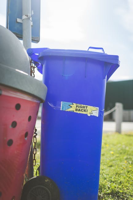 A large green waste bin filled with a mix of cardboard boxes, some flattened and others partially open, along with white packaging containers and small paper scraps. The surface of the bin shows signs of wear with scratches and graffiti. Leaning against the bin are wooden pallets and flat-pack furniture components, including a panel with a reflective surface. In the background, a green waste collection truck with a hydraulic lift mechanism is parked on an urban street, surrounded by trees, multi-storey buildings, and a clear sky. The scene appears to represent a private rubbish collection or on-site clearance, with the waste prepared for removal by local waste management services such as Waste Collection Fulham. The environment suggests a typical urban setting where rubbish is temporarily staged prior to disposal or recycling, emphasizing the practicalities of alternative waste handling methods in Fulham.